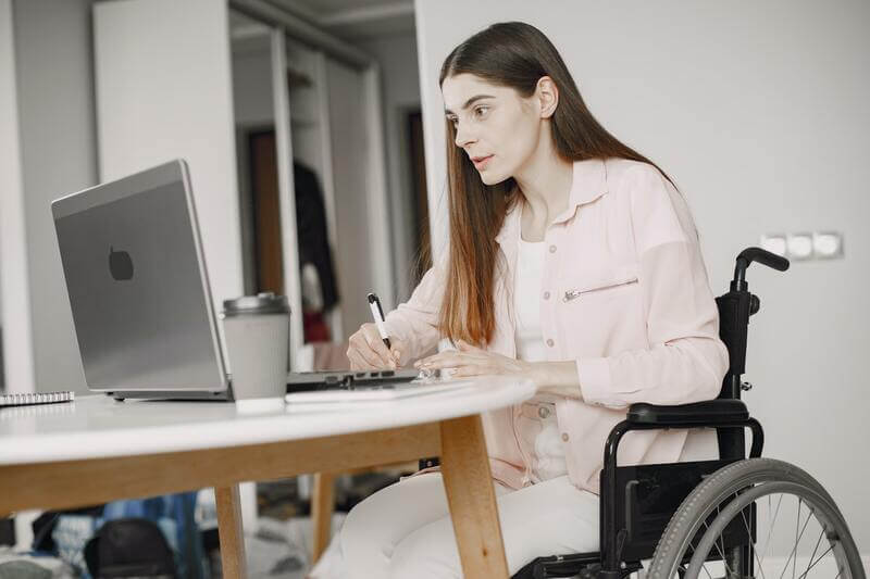 Mulher sentada em uma cadeira de rodas e segurando uma caneta enquanto olha para um notebook sobre uma mesa.